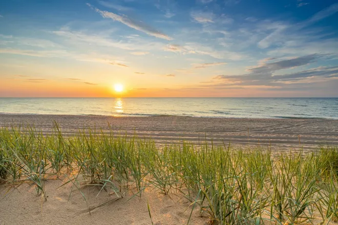 Le Soleil sur l’horizon marin, vu d’une plage avec des herbes dunaires au premier plan.